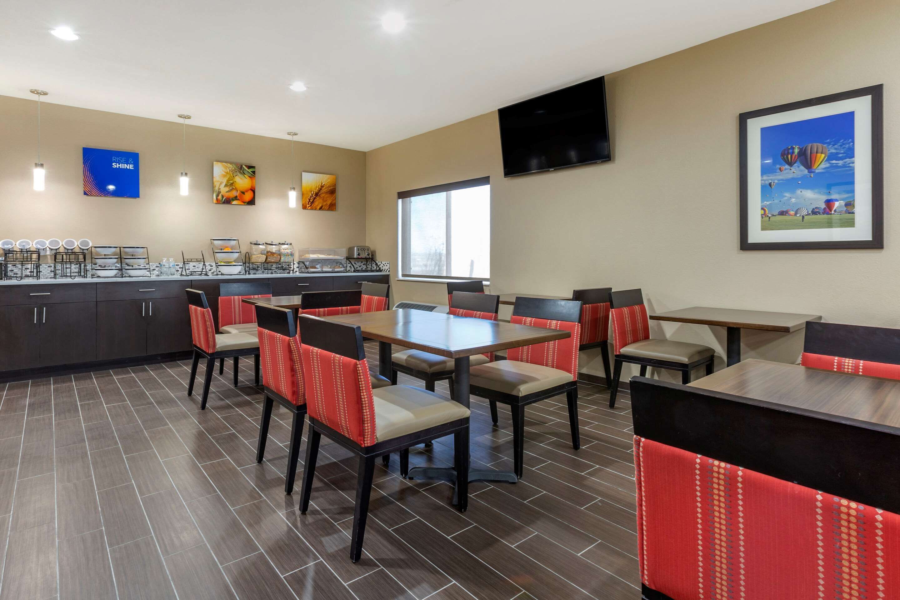 A hotel dining area with multiple tables and chairs, a kitchen area, and a large window providing natural light.