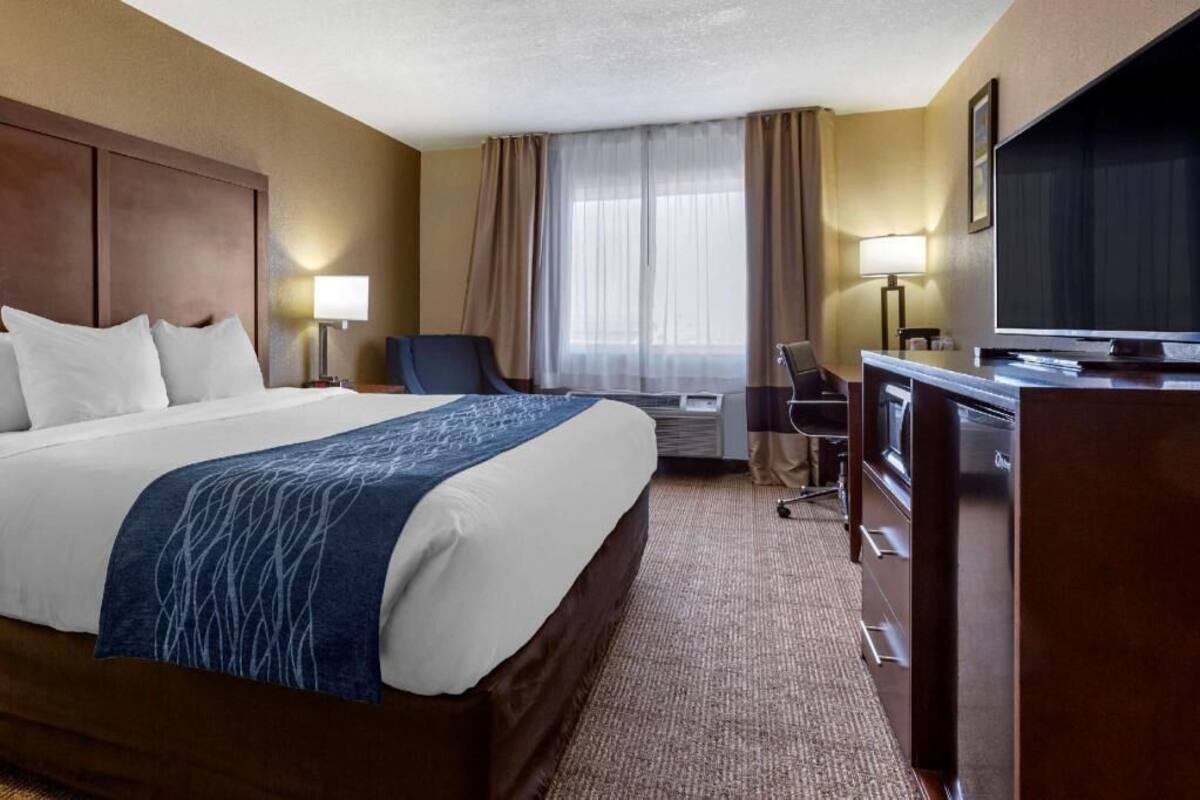 A hotel room with a large bed, a desk with a television, a chair, and a desk lamp.