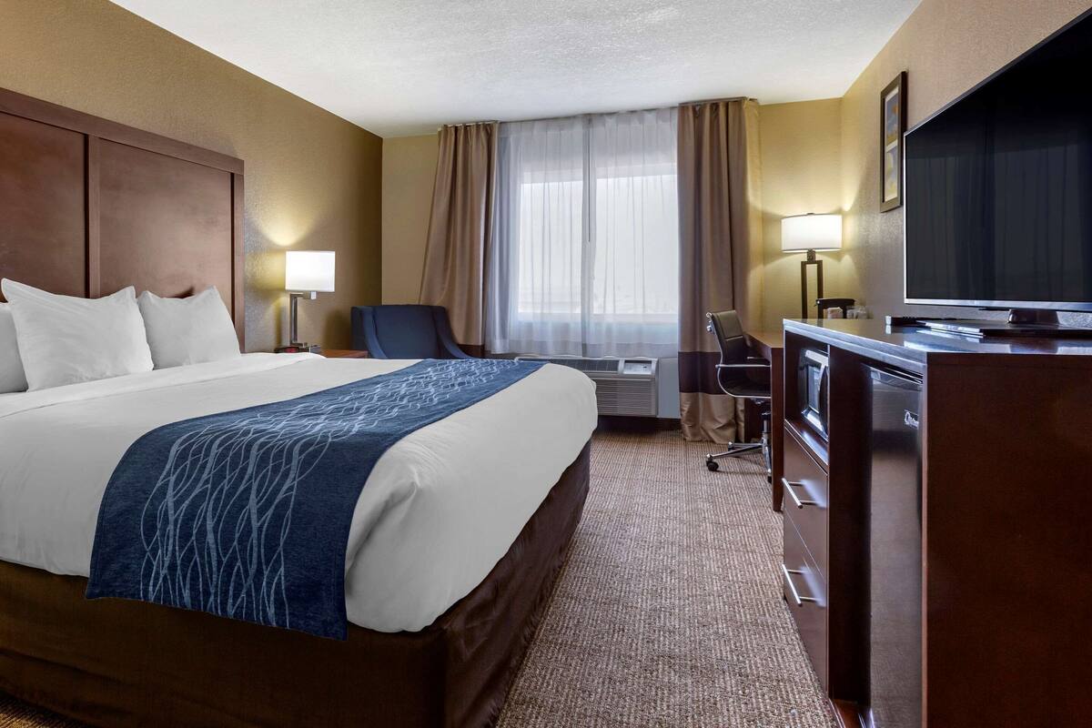 A hotel room with a large bed, a desk with a television, a chair, and a desk lamp.