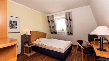 Quarto de hotel com cama, escrivaninha, cadeira, mesa de cabeceira, abajur, quadro emoldurado e janela com cortinas.