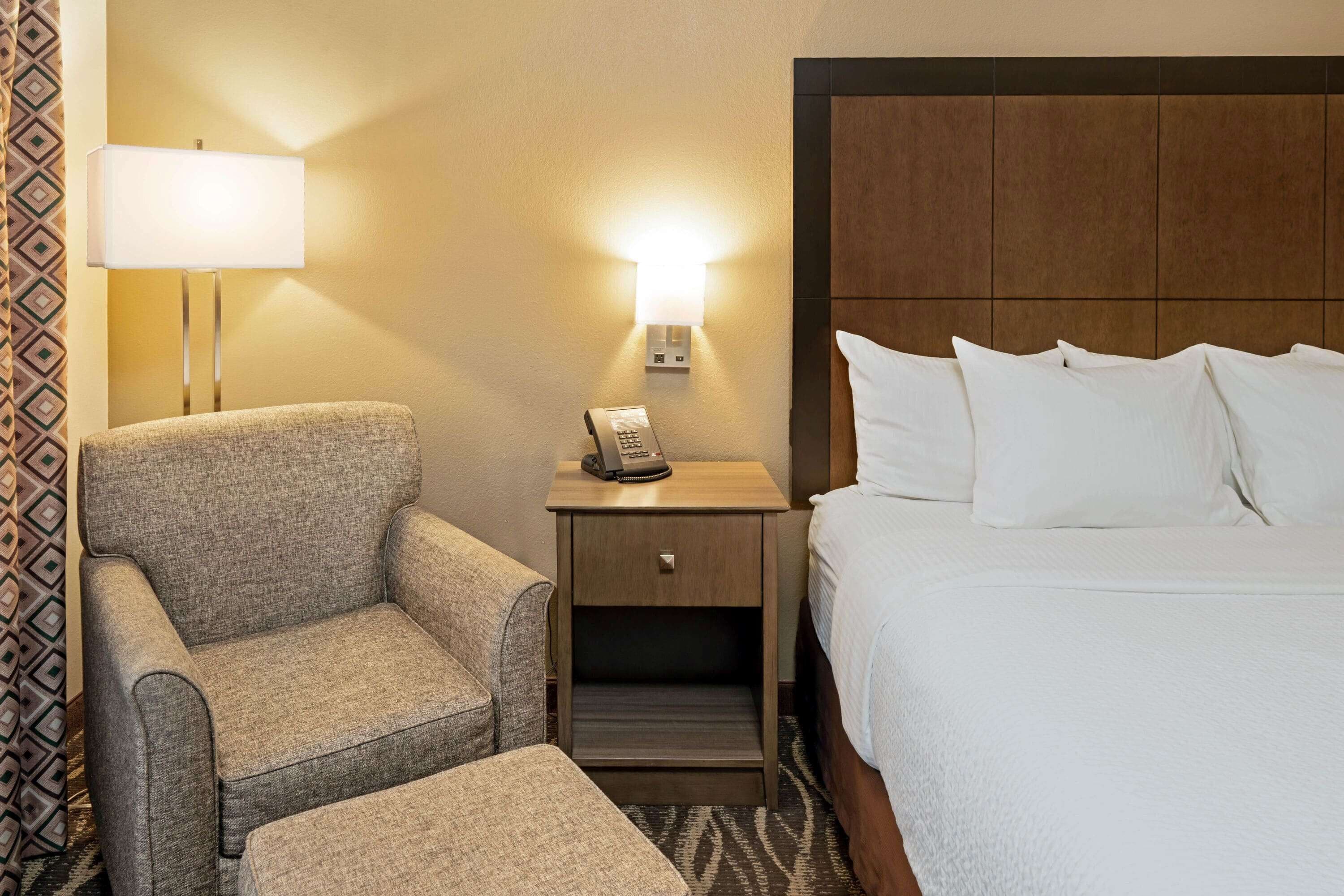 A hotel room with a bed, a nightstand with a lamp, a telephone, and a chair.