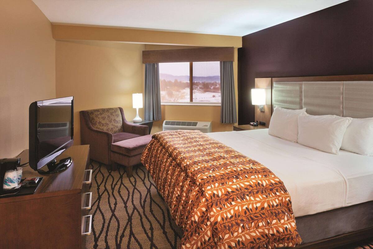 A hotel room with a bed, a TV, a desk with a lamp, a chair, and a window with curtains.