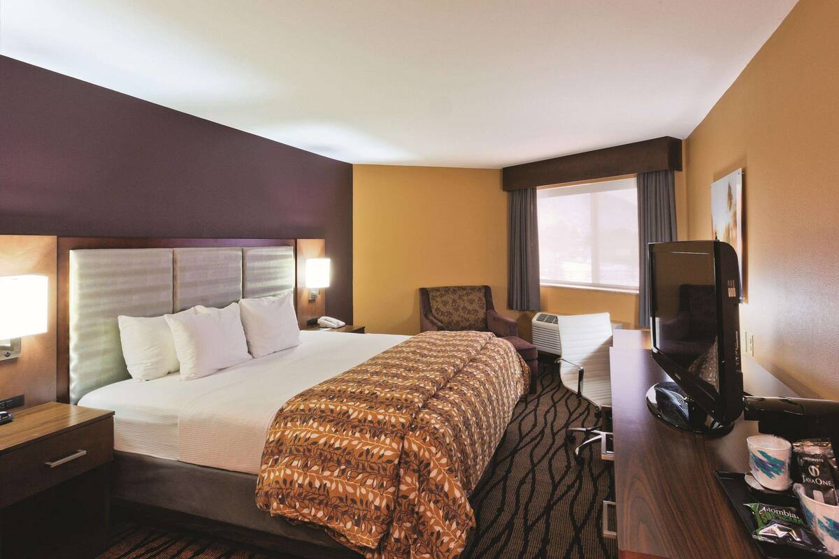A hotel room with a large bed, a desk with a computer, a TV, and a window with curtains.