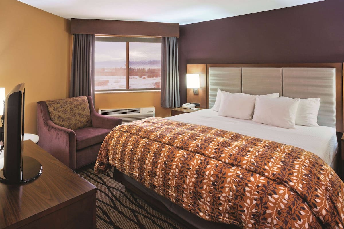 A hotel room with a large bed, a desk with a flat-screen TV, a chair, an air conditioning unit, and a window with a view of a snowy landscape.