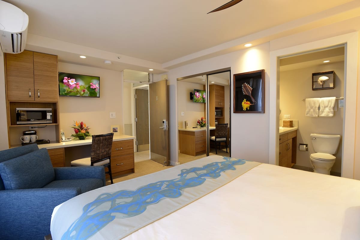 Habitación de hotel con cama, escritorio, silla, televisor, espejo, baño y kitchenette.