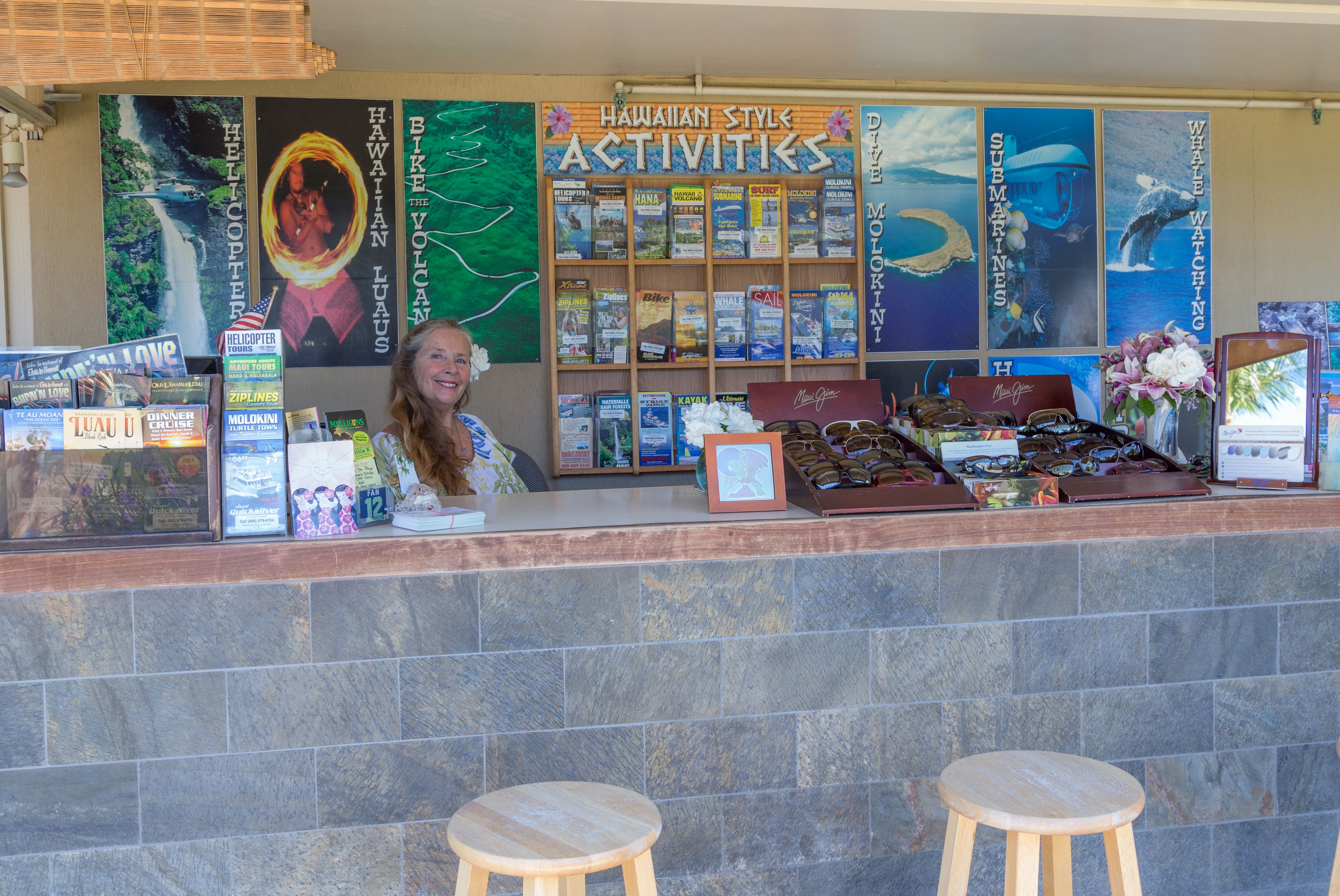 A counter with a display of travel books and souvenirs, a signboard with 'Hawaiian Style Activities', and a woman standing behind the counter.