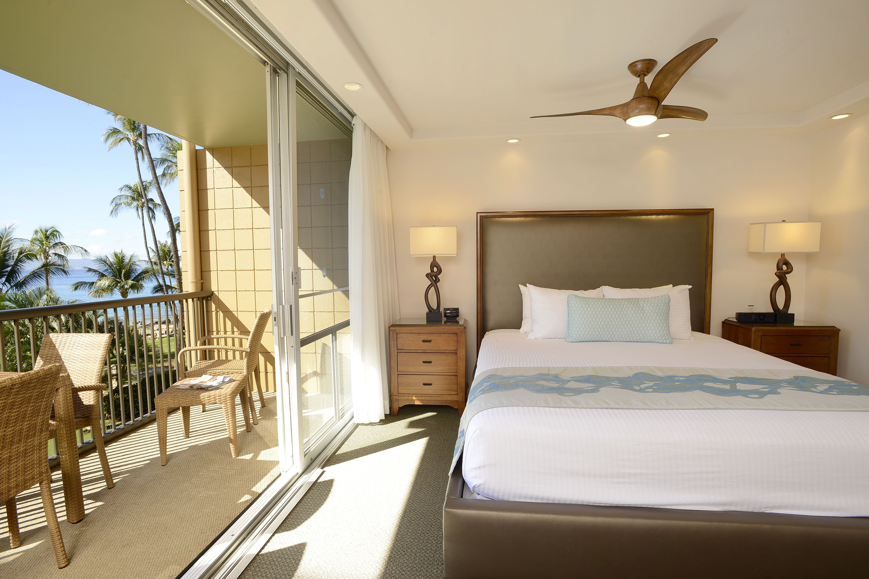 A hotel room with a balcony, a bed with a headboard, two bedside tables with lamps, and a ceiling fan.