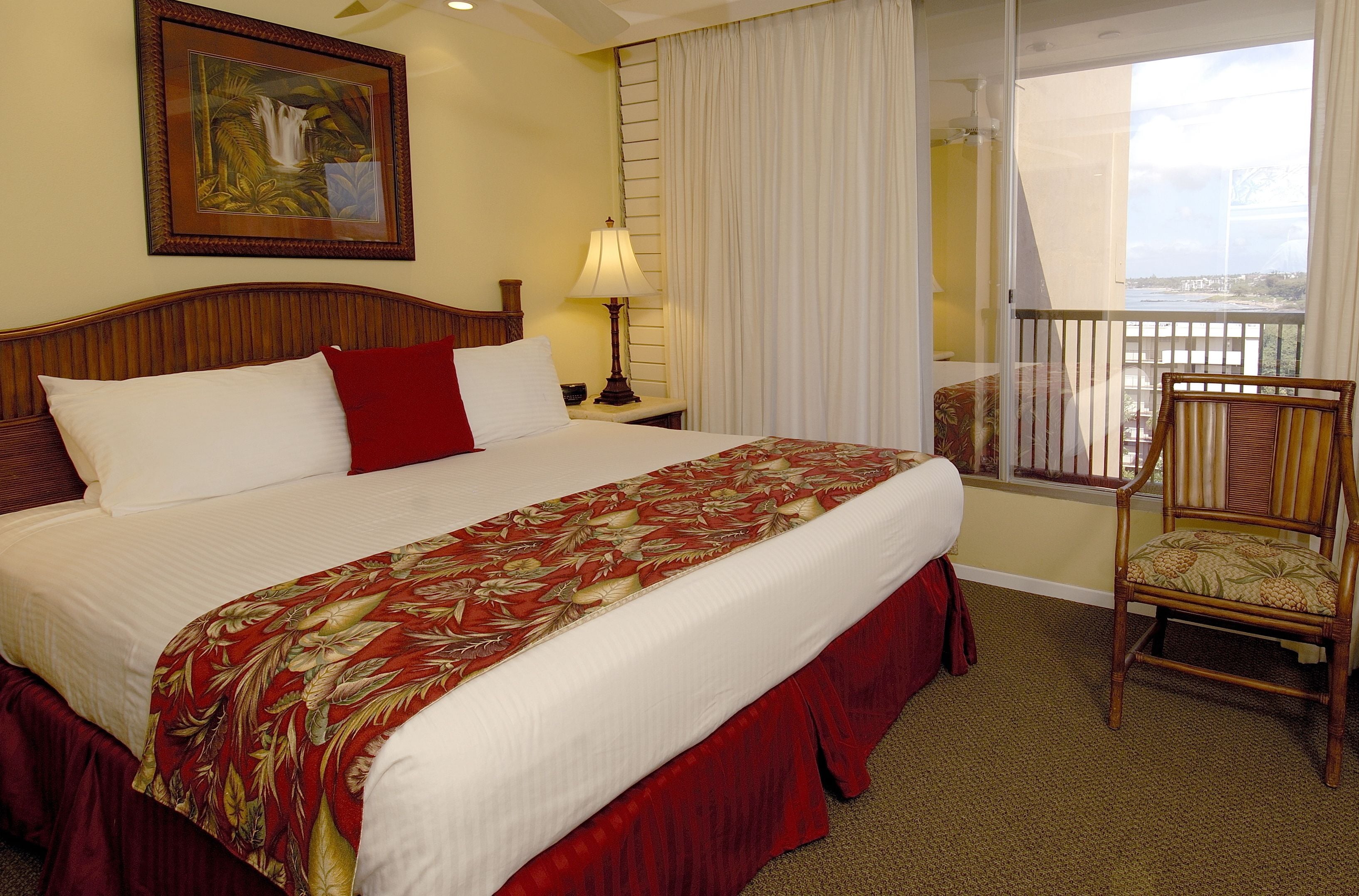 Habitación de hotel con una cama grande, una silla, una mesita de noche con lámpara, un cuadro en la pared y un balcón con vistas.