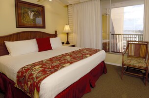 Habitación de hotel con una cama grande, una silla, una mesita de noche con lámpara, un cuadro en la pared y un balcón con vistas.