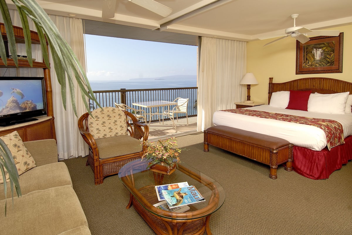 Habitación de hotel con cama, televisor, mesa de comedor y balcón con vista al océano.