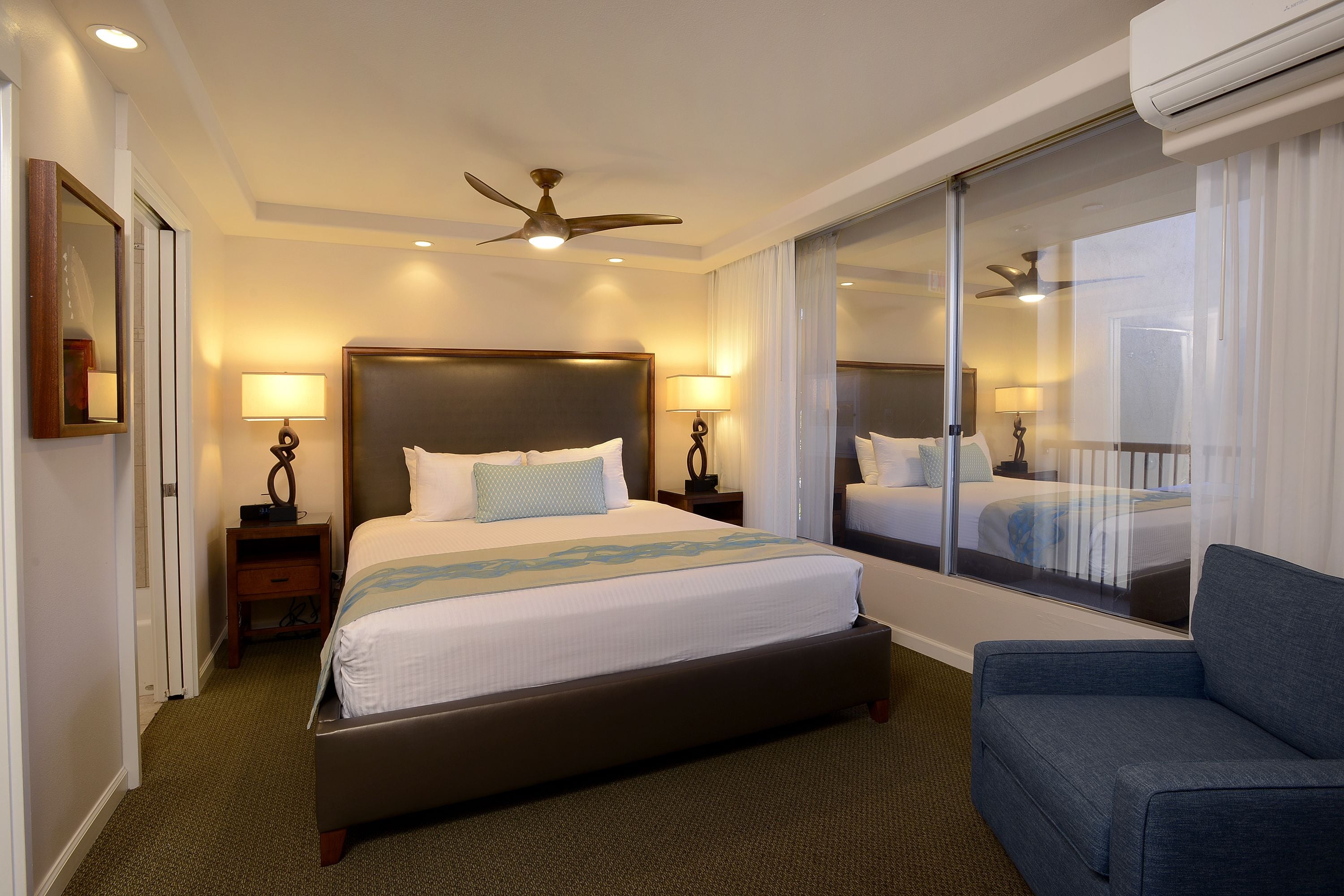 A hotel room with a large bed, two bedside tables with lamps, a ceiling fan, and a large window with curtains.