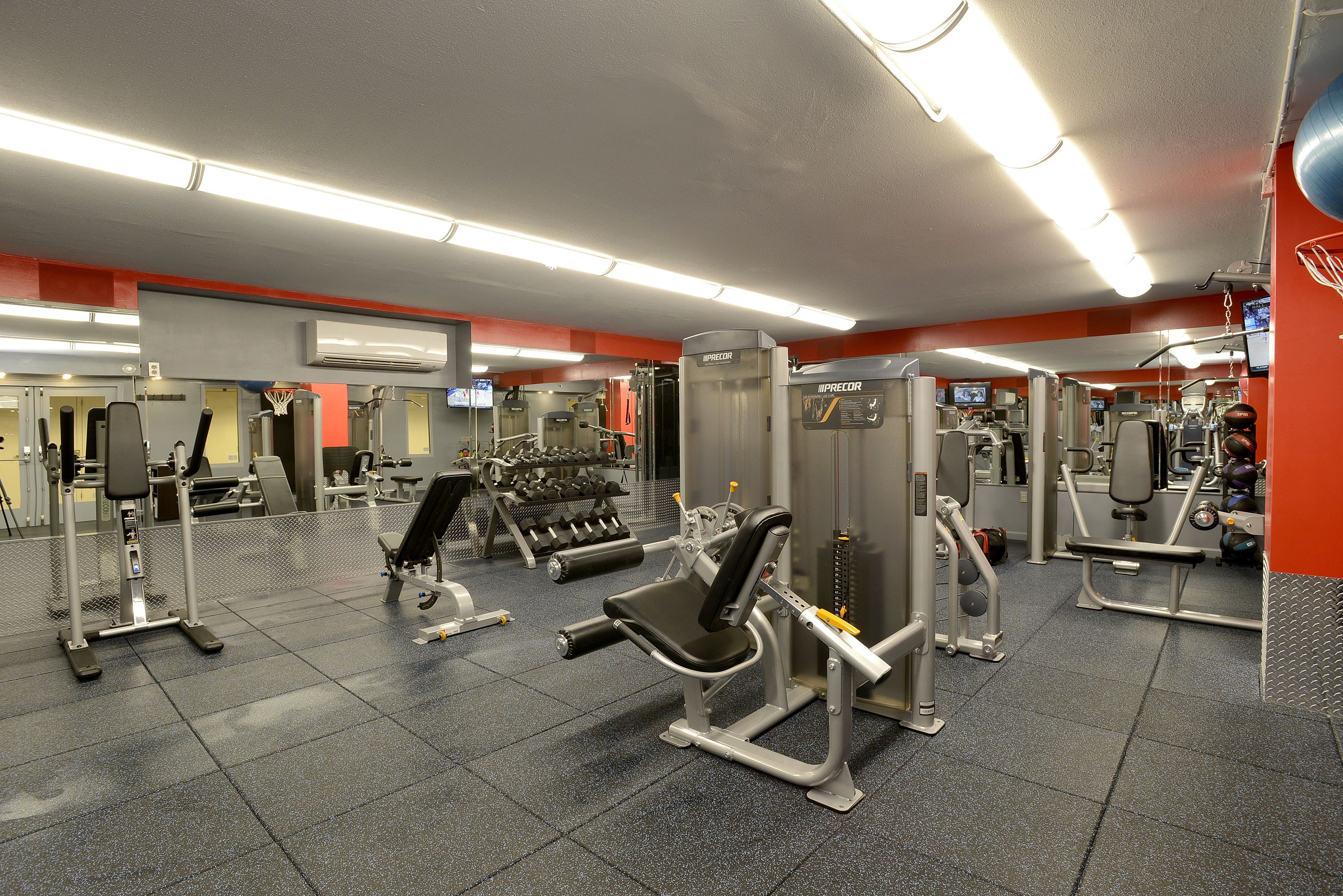 A gym with various exercise machines, a basketball hoop, and a mirrored wall.