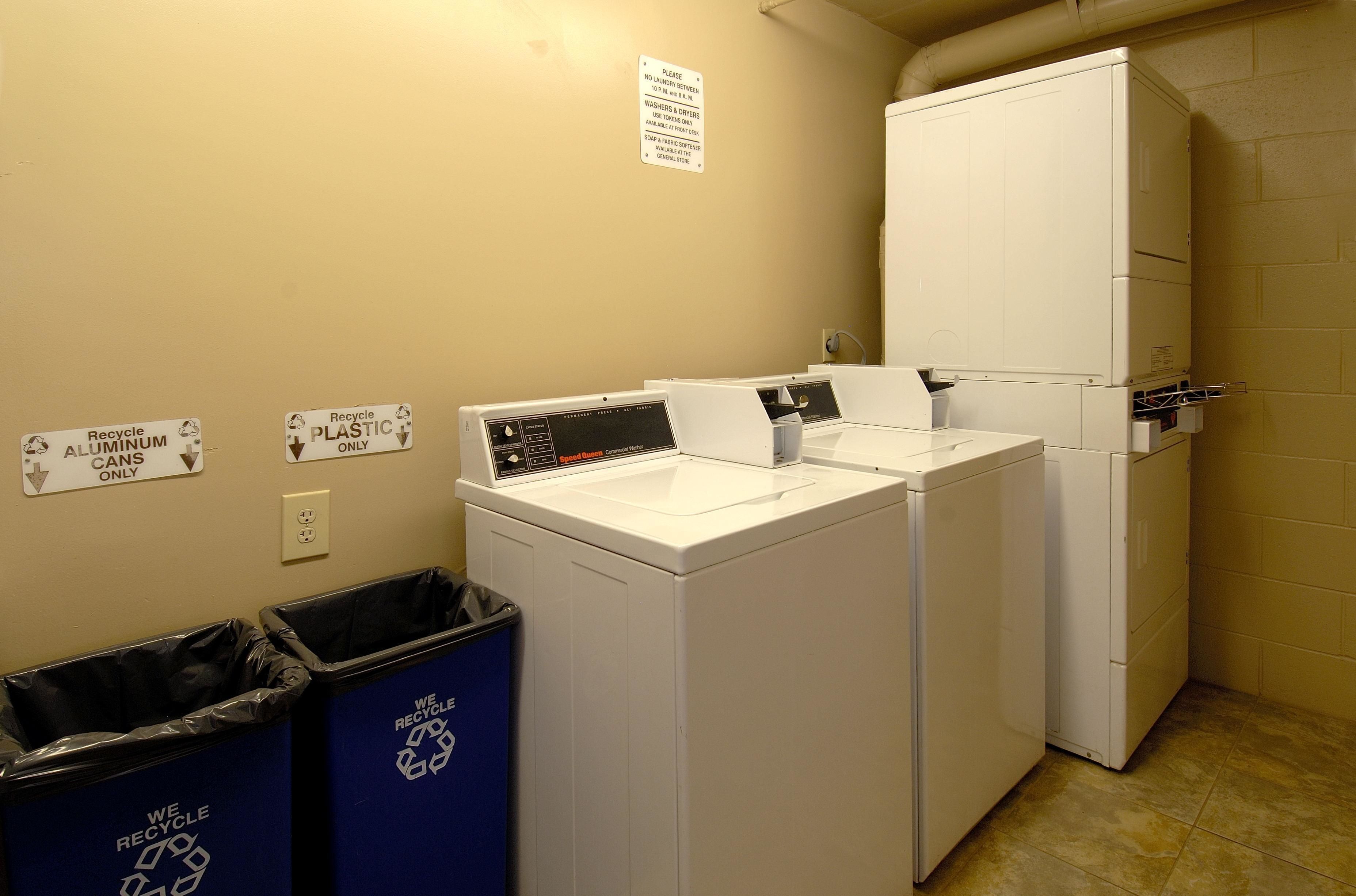 A laundry area with two washing machines, a dryer, recycling bins, and a sign indicating recycling instructions.