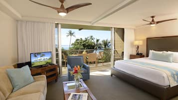A hotel room with a bed, a sofa, a coffee table with a magazine, a TV, and a balcony with a view of palm trees and the ocean.