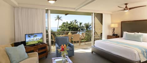 Ein Hotelzimmer mit Bett, Sofa, Couchtisch mit Zeitschrift, Fernseher und Balkon mit Blick auf Palmen und Meer.