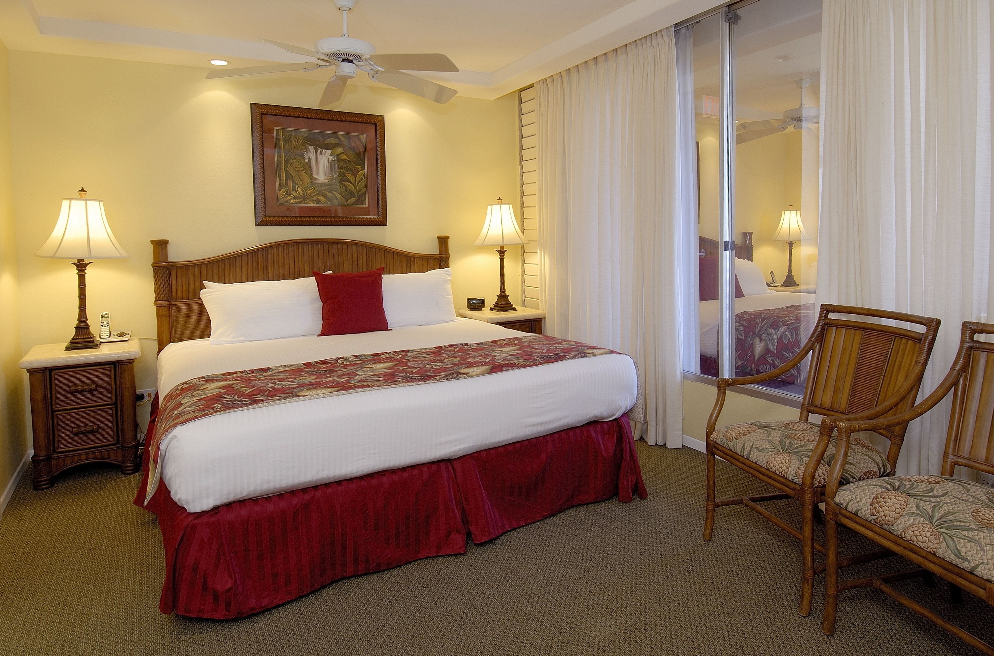 Una habitación de hotel con una cama grande, dos mesitas de noche con lámparas, una silla de madera, un ventilador de techo y un cuadro en la pared.