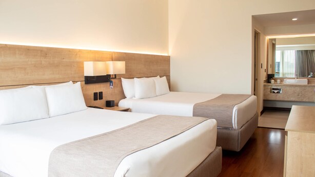 Une chambre d’hôtel avec deux lits, une tête de lit en bois, des lampes de chevet, une table de chevet, un petit bureau, un miroir et une salle de bain avec un lavabo et un miroir.