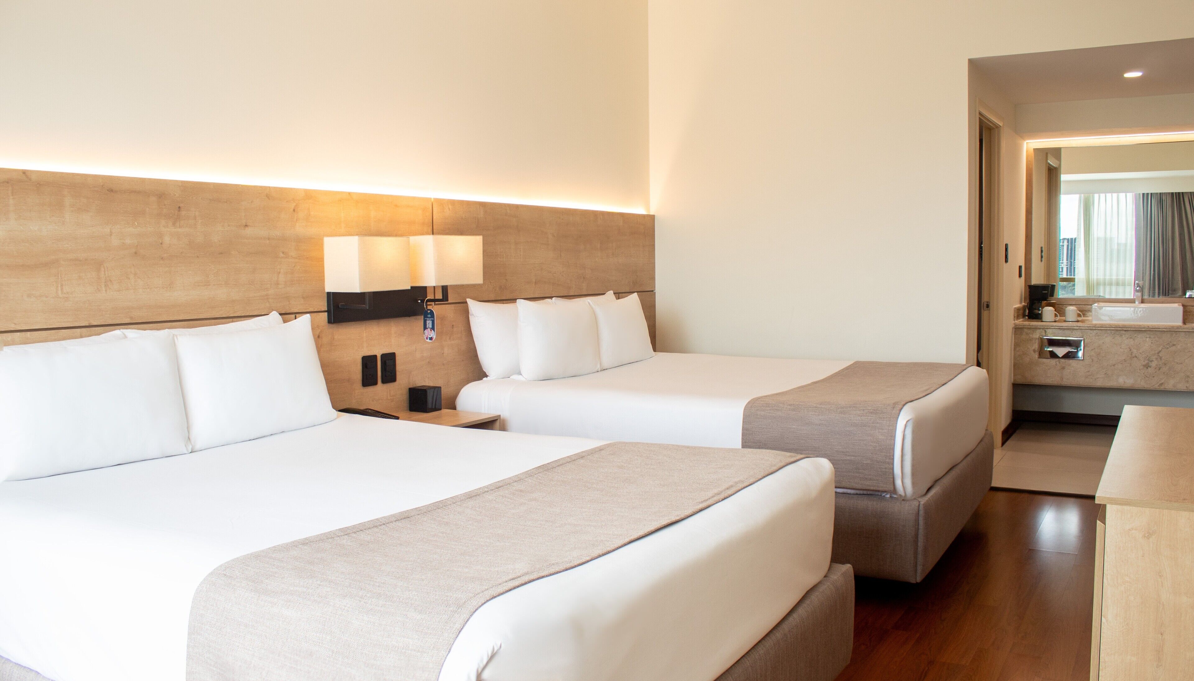 A hotel room with two beds, a wooden headboard, bedside lamps, a bedside table, and a small seating area with a chair.