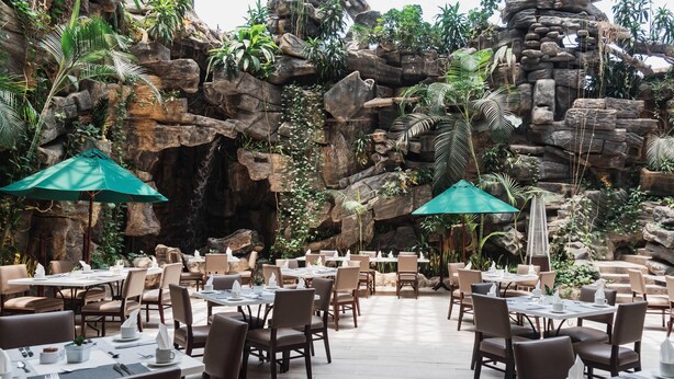Un restaurante con mesas y sillas dispuestas en un espacio al aire libre, con un telón de fondo rocoso que recuerda a una cueva.