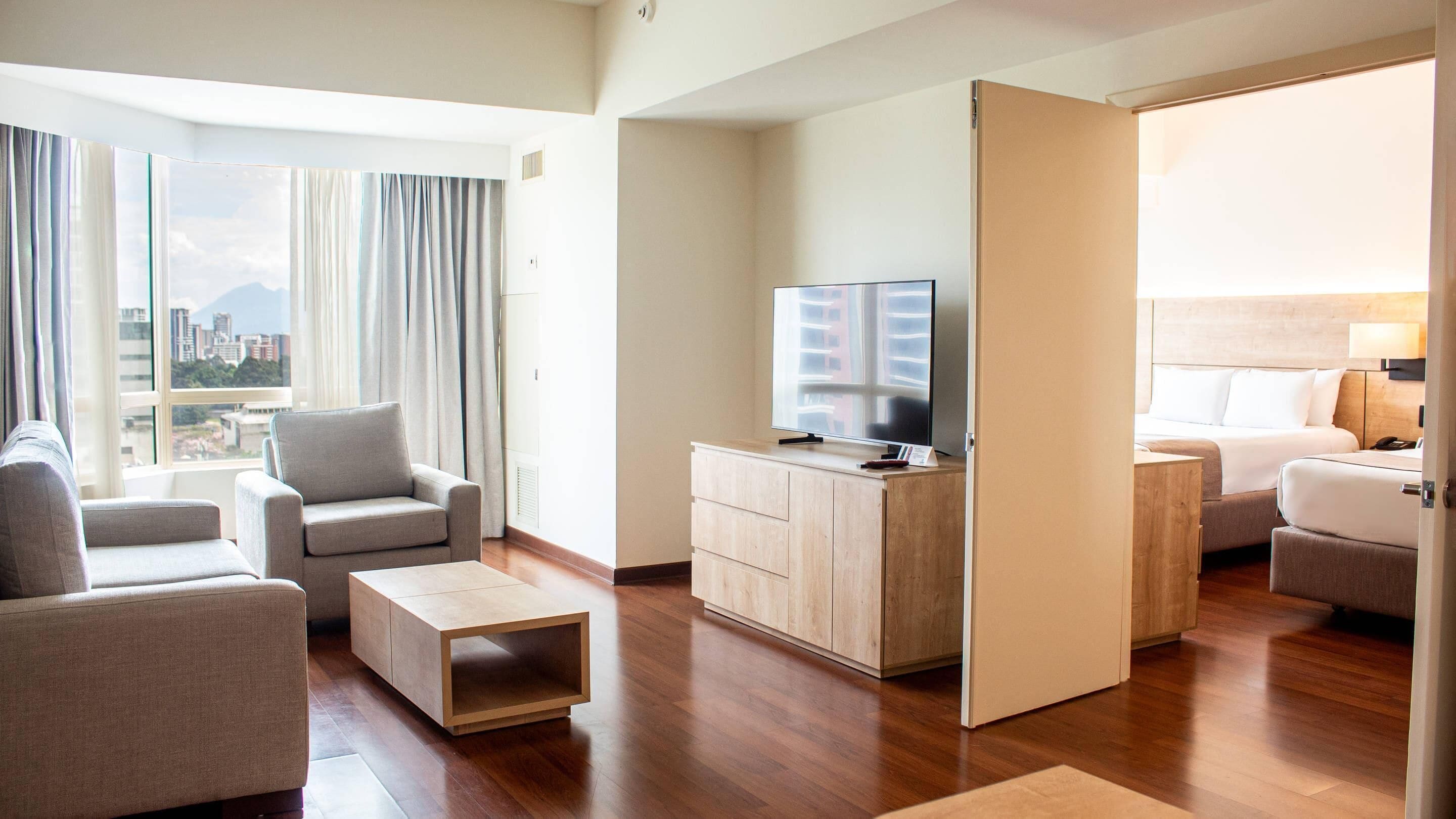 Una habitación de hotel moderna con sofá, una mesa de centro, televisión, cama y vistas a la ciudad.