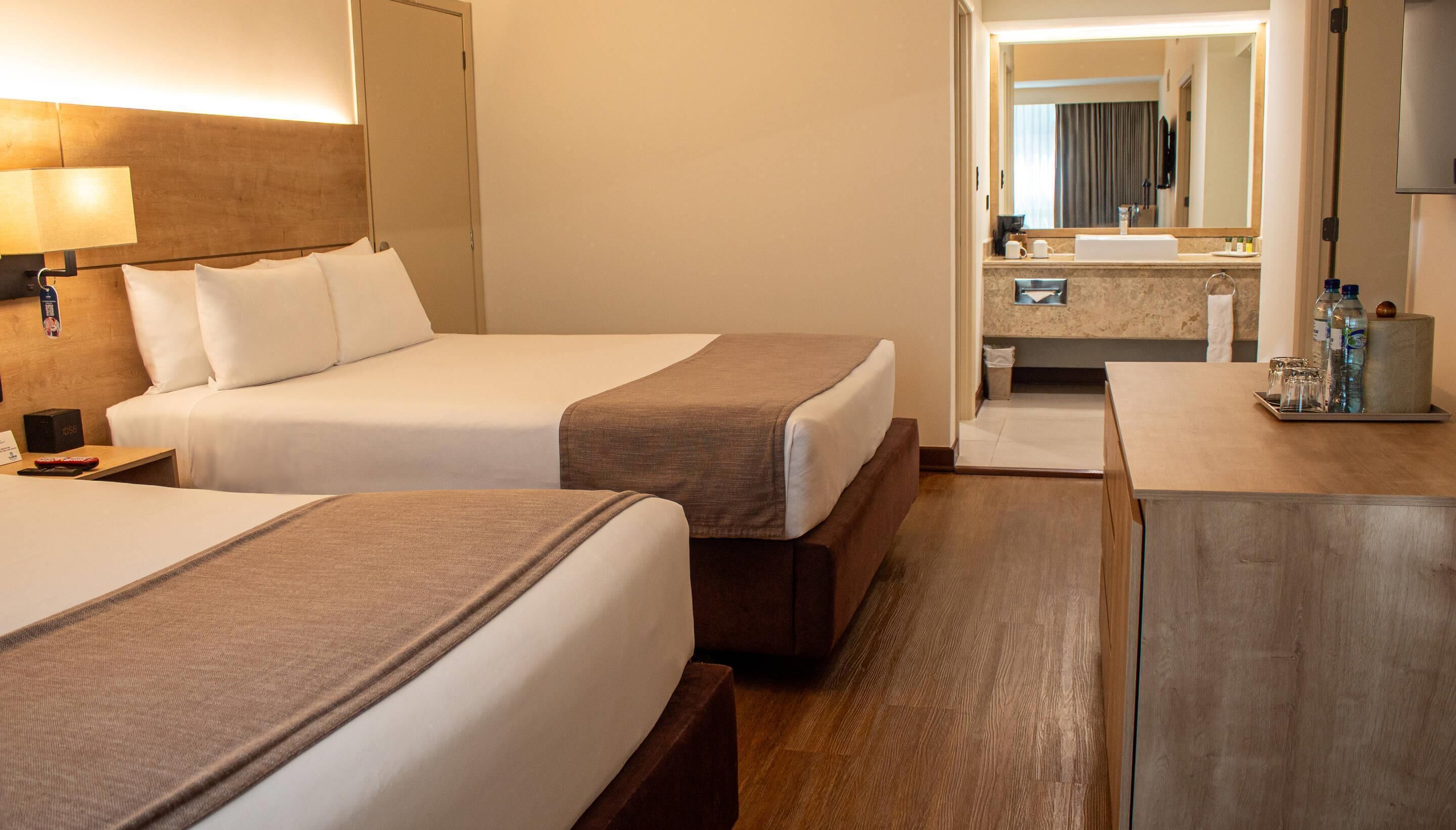 A hotel room with two beds, a bedside table with a lamp, a bathroom with a sink and mirror, and a small cabinet with toiletries.