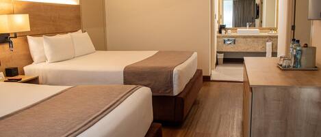 A hotel room with two beds, a bedside table with a lamp, a bathroom with a sink and mirror, and a small cabinet with toiletries.