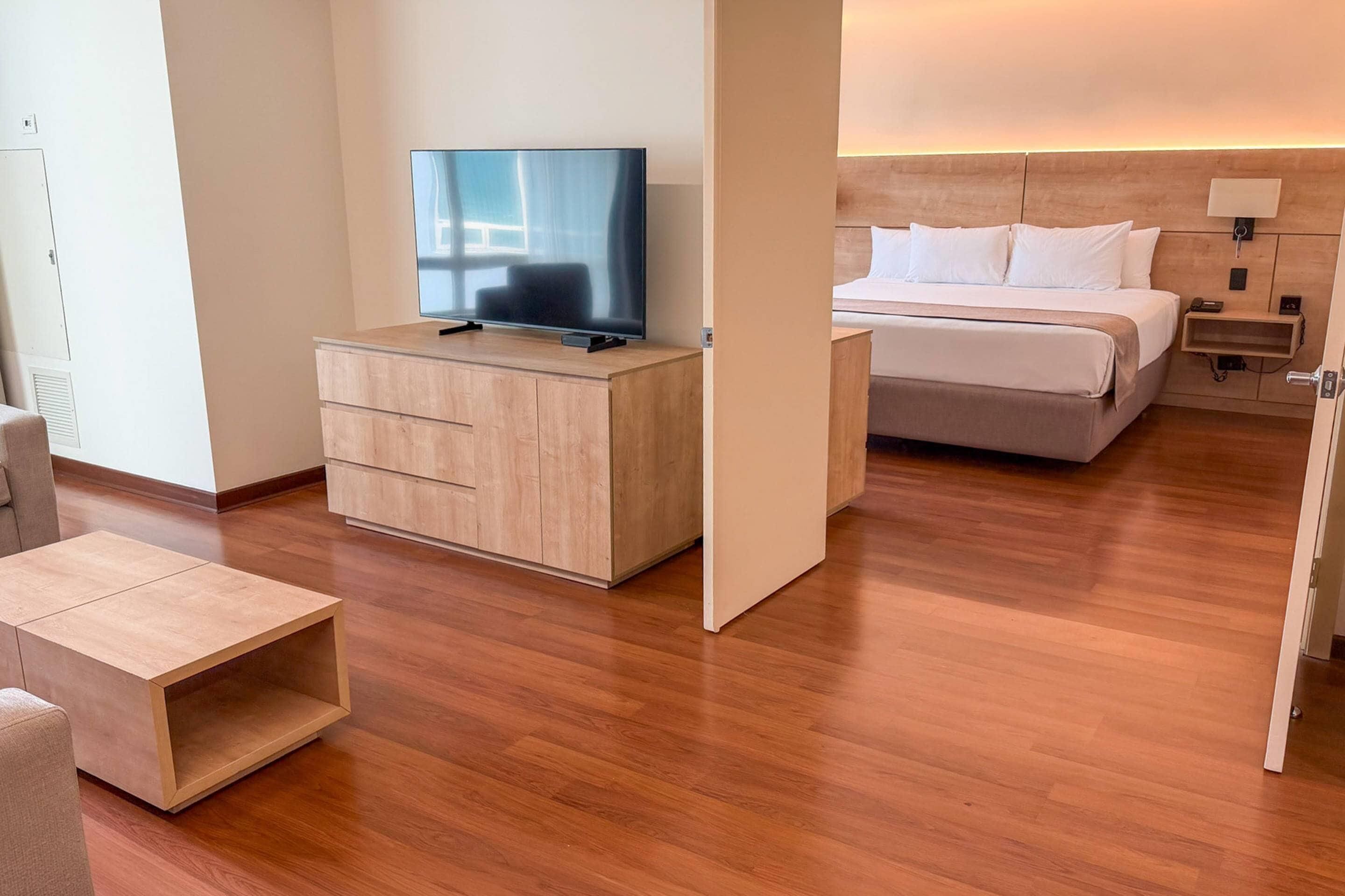 A modern hotel room with a wooden floor, a bed with white bedding, a TV on a wooden cabinet, and a bedside table with a lamp.