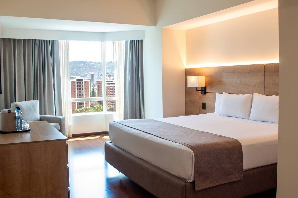 Habitación de hotel con una cama grande, un escritorio, una lámpara y vistas a edificios a través de la ventana.