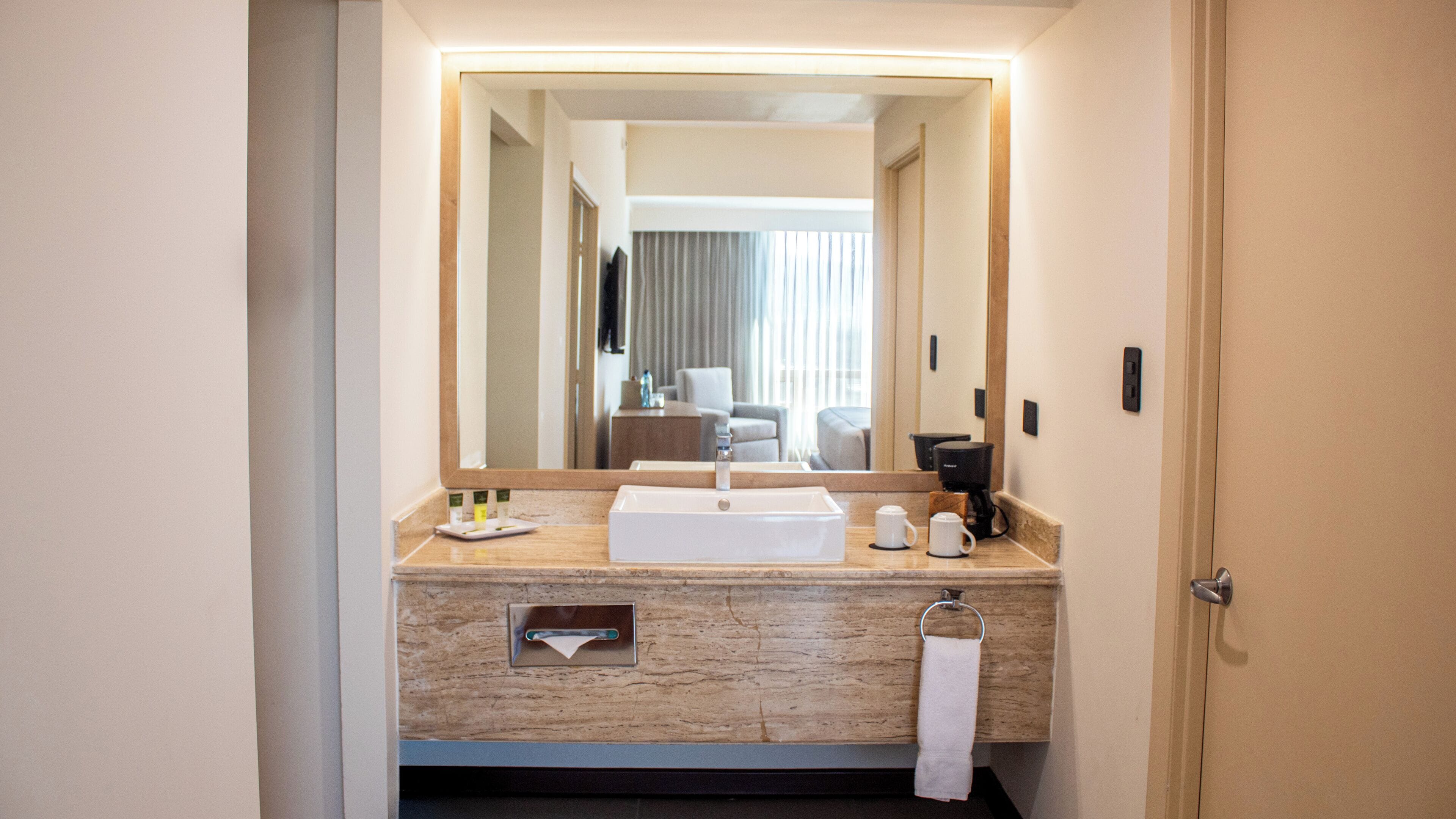 Bagno d'albergo con uno specchio grande, un lavandino, un portascopevoli e una macchina per il caffè.