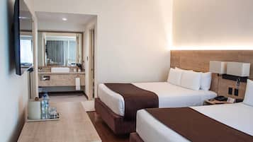 Habitación de hotel con dos camas, un televisor de pantalla plana, una mesita de noche con lámpara, y un baño visible a través de una puerta abierta.