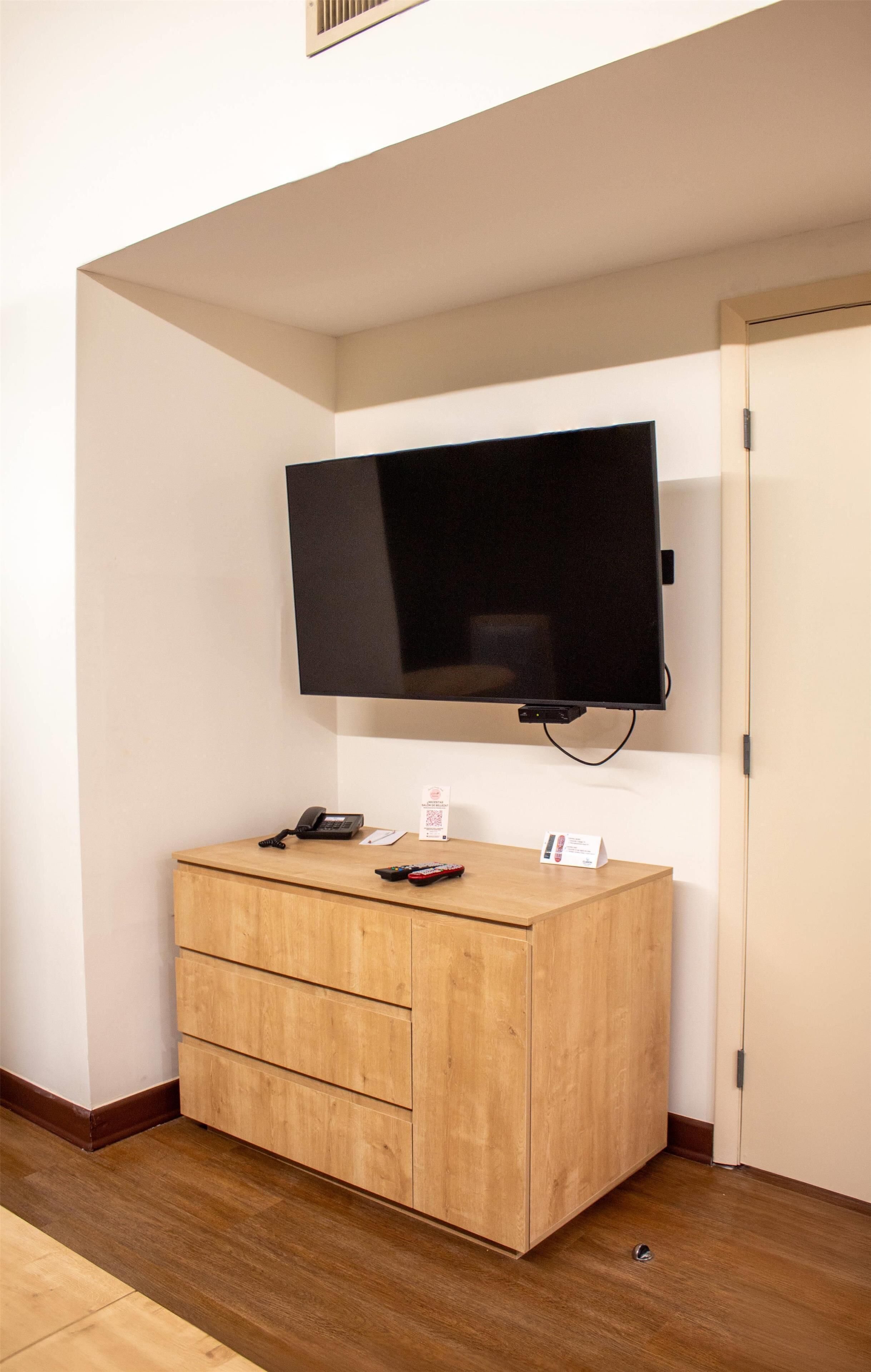 Quarto com televisão de tela plana na parede, um armário de madeira com gavetas, um telefone e uma pequena placa no armário.