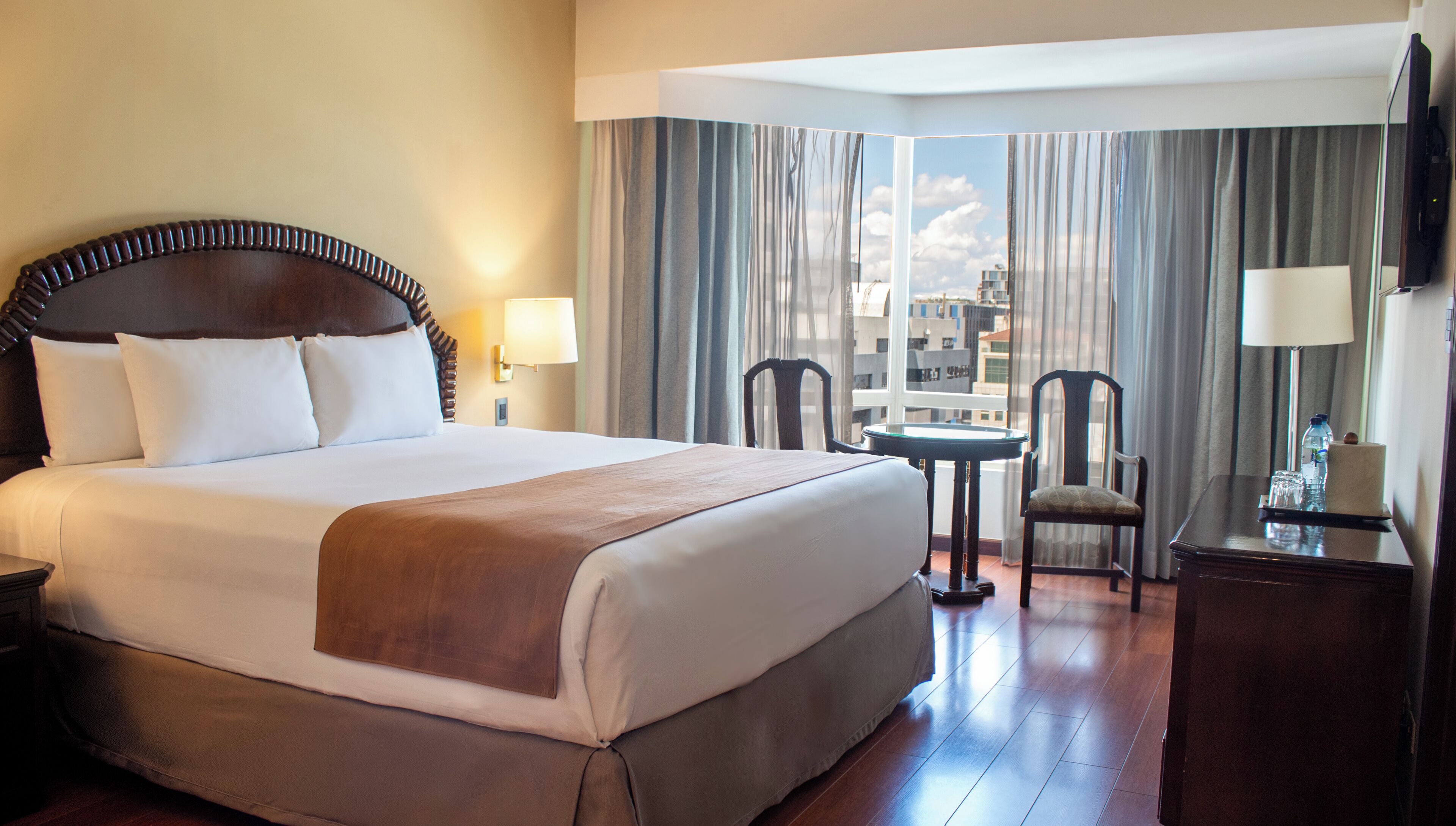 Habitación de hotel con una cama grande, un escritorio con silla, ventana con cortinas y vista a edificios.