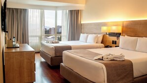 A hotel room with two beds, a desk, a TV, and a view of the city.