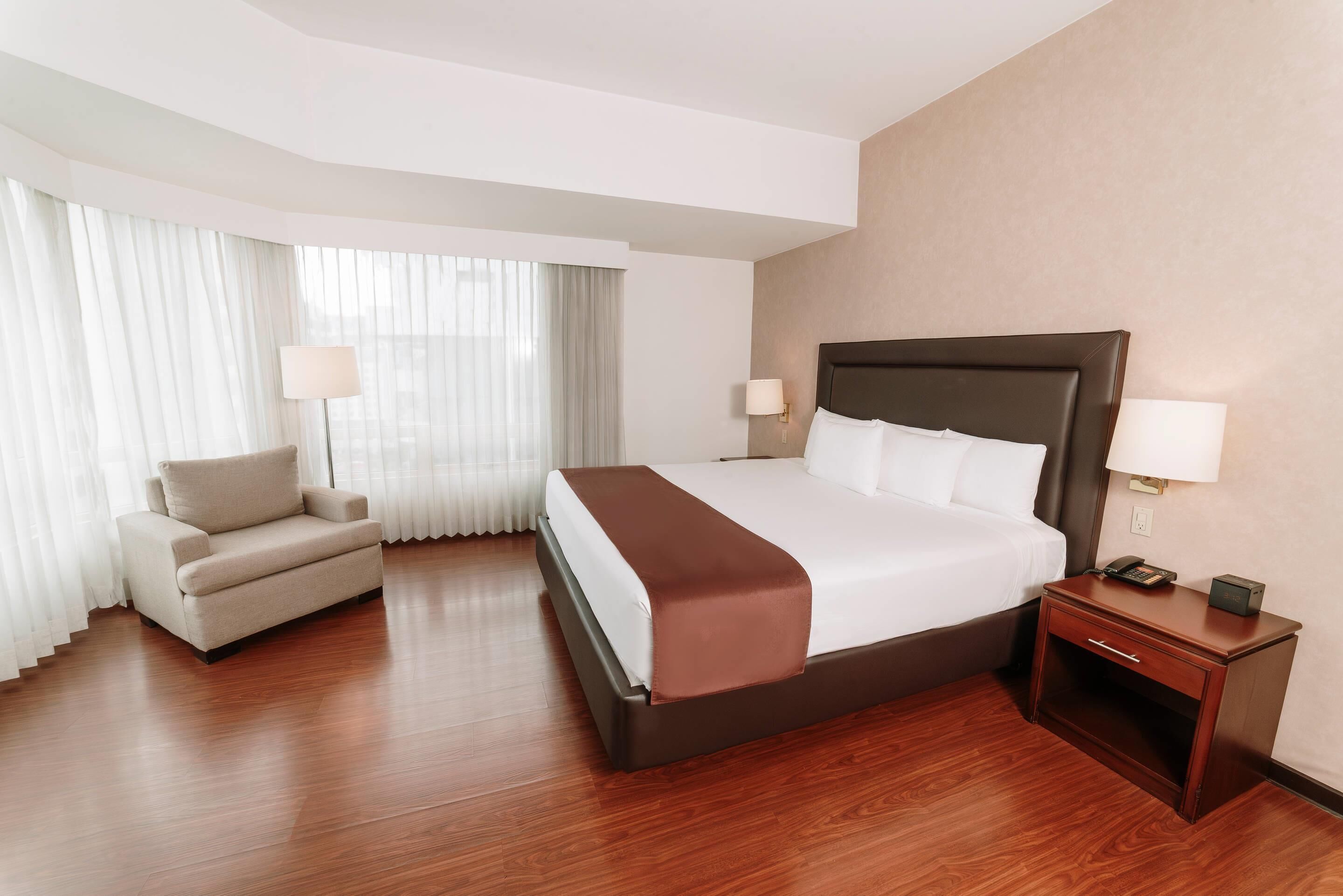 Habitación de hotel con cama, mesita de noche, lámpara, silla y ventana grande con cortinas.