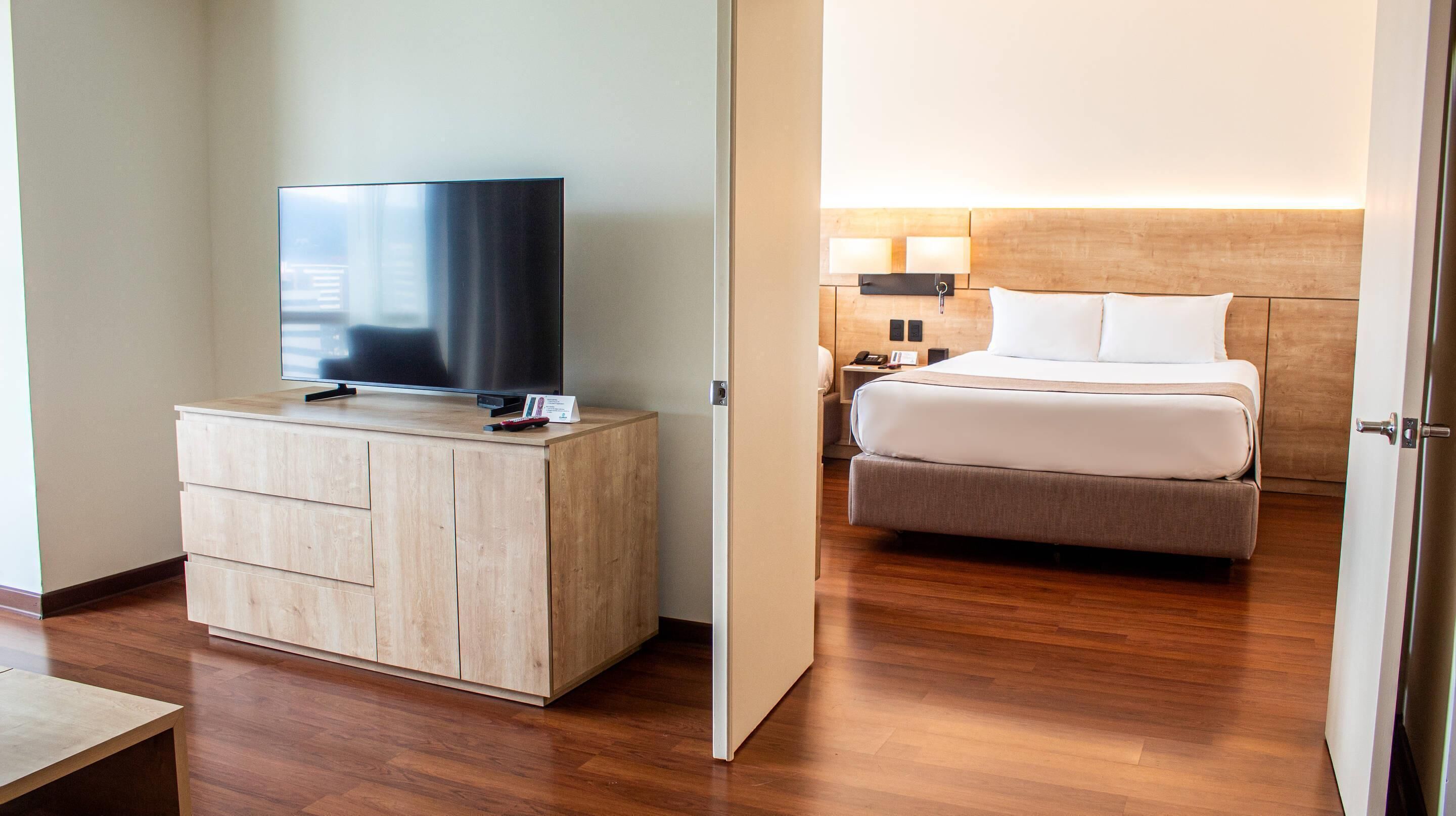 A hotel room with a wooden dresser, a flat-screen TV, a bed with white bedding, a nightstand, and a wall-mounted lamp.