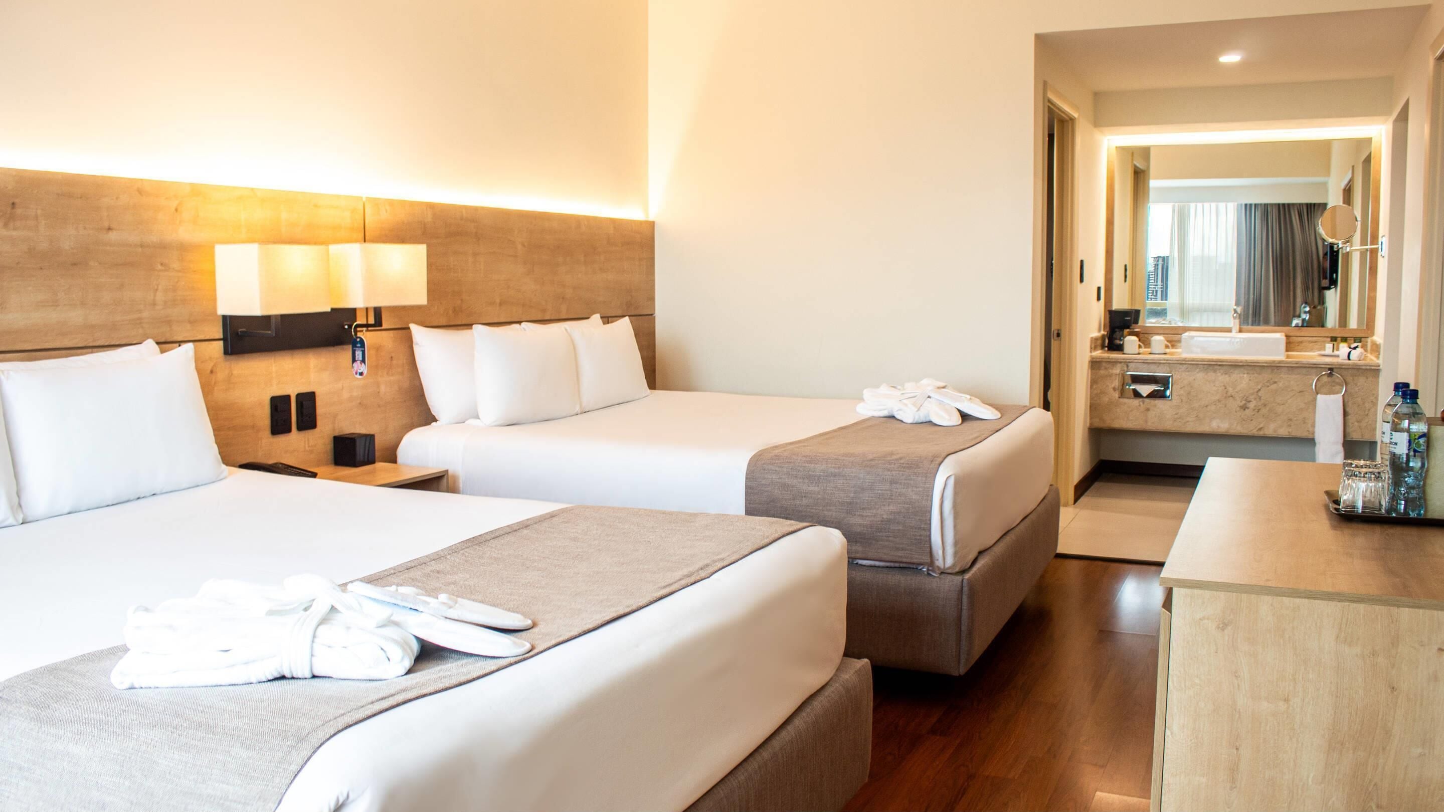 A hotel room with two beds, a bedside table, a desk, a mirror, and a bathroom visible through an open door.