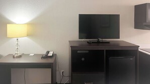Habitación con escritorio, lámpara, televisor de pantalla plana, refrigerador y mueble de pared.