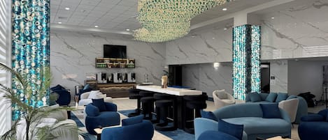 A modern hotel lobby with blue sofas, a bar area, and a decorative chandelier.