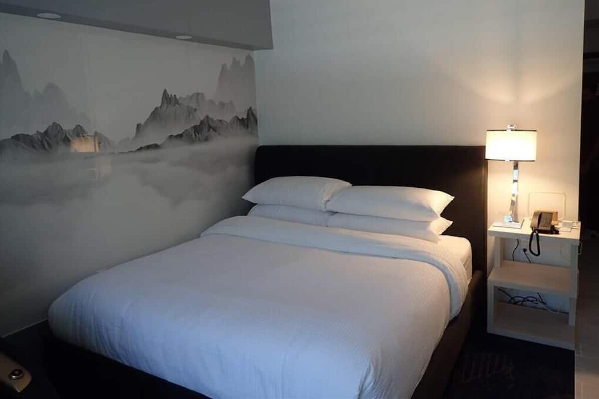Uma cama bem arrumada com roupa de cama branca, uma luminária na parede e uma mesa de cabeceira com um telefone e carregador.