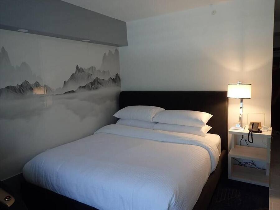 A neatly made bed with white linens, a wall-mounted lamp, and a nightstand with a phone and charger.