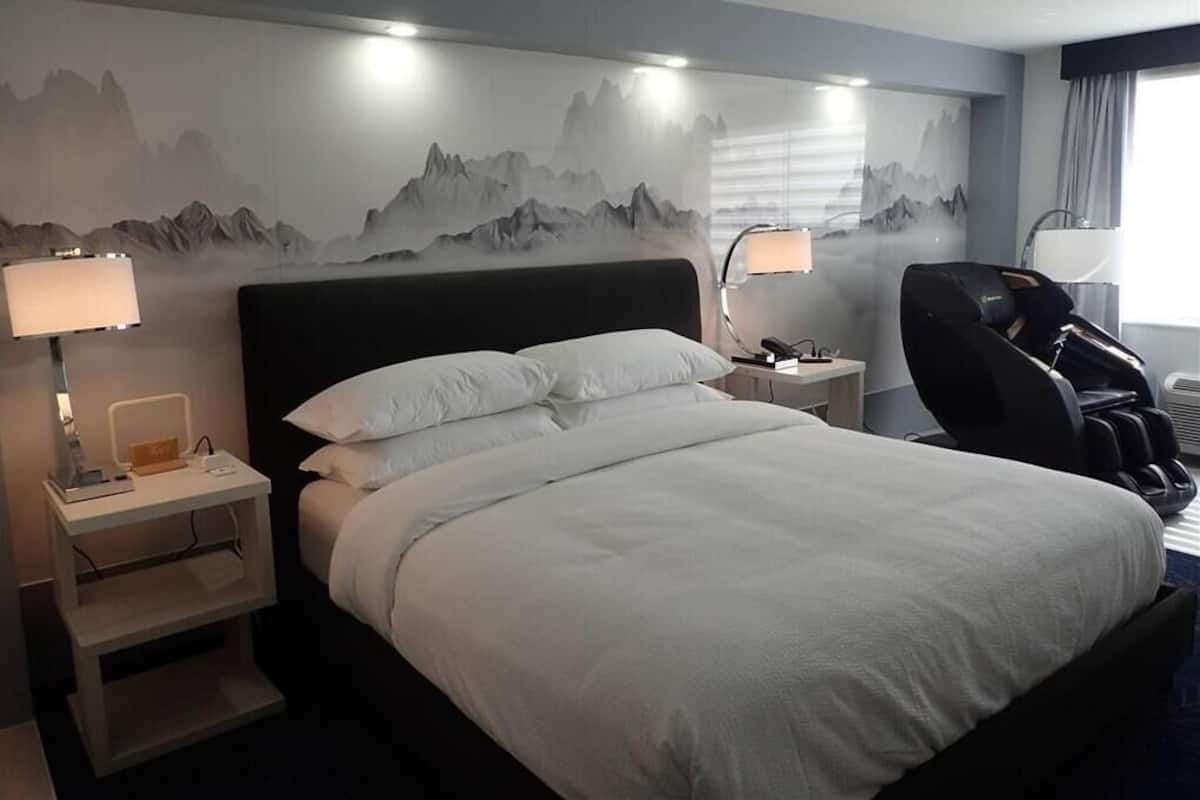 Quarto de hotel com uma cama grande, duas mesas de cabeceira com luminárias, uma cadeira de massagem e um mural de montanhas na parede.