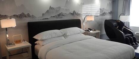 Una habitación de hotel con una cama grande, dos mesitas de noche con lámparas, una silla de masajes y un mural de montañas en la pared.