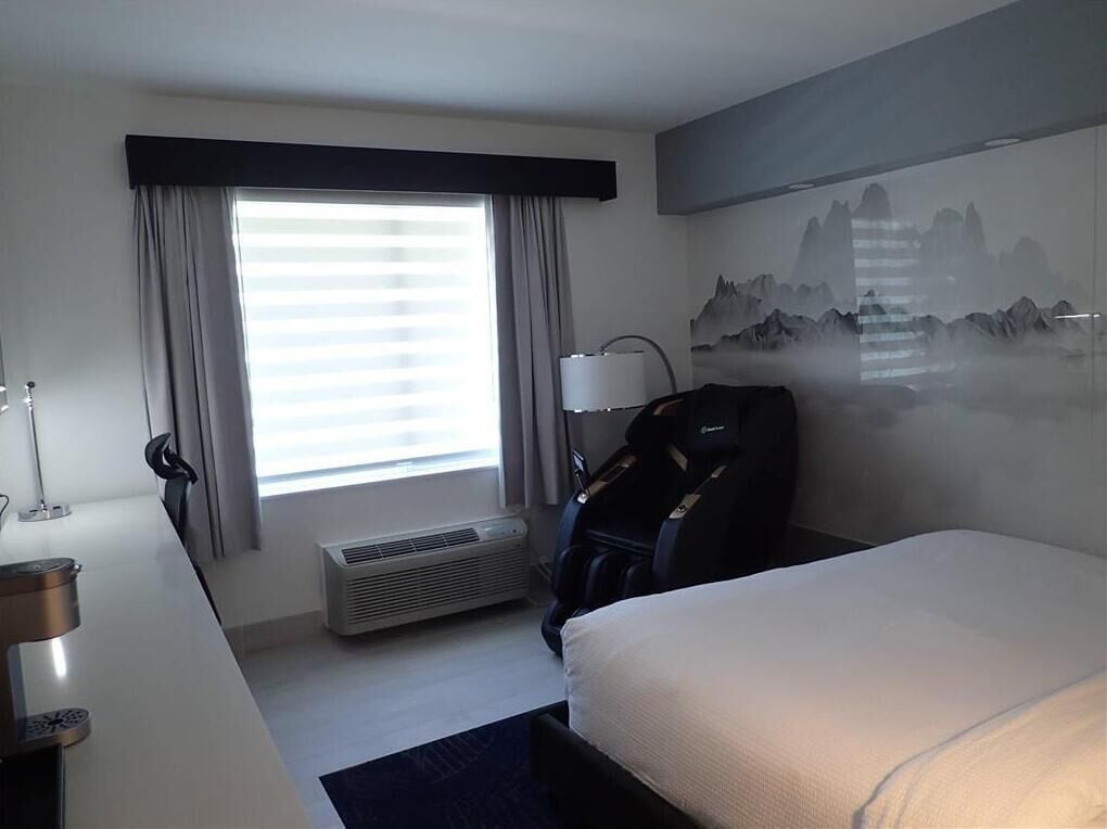 A hotel room with a bed, a desk, a chair, a window with curtains, a wall-mounted air conditioner, and a painting of mountains.