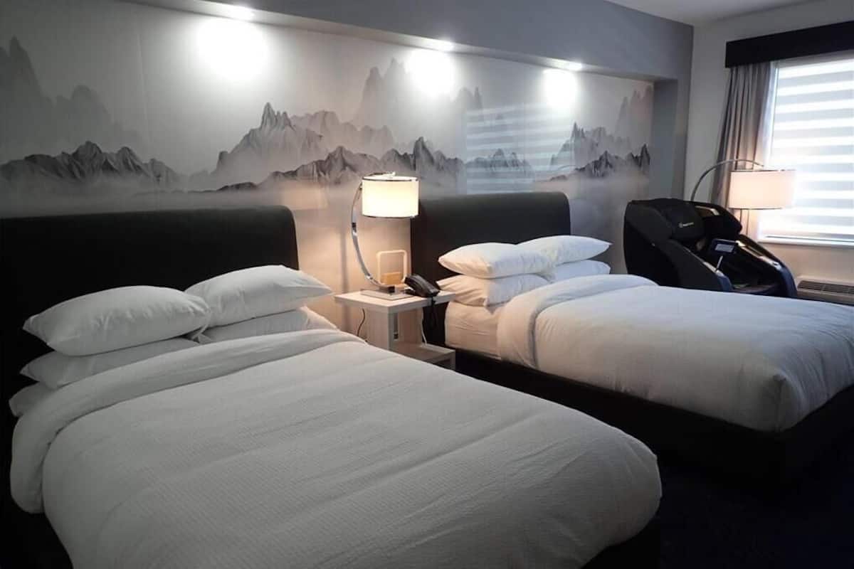 Quarto de hotel com duas camas, uma cadeira de massagem, uma mesa de cabeceira com abajur e um mural de montanhas na parede.