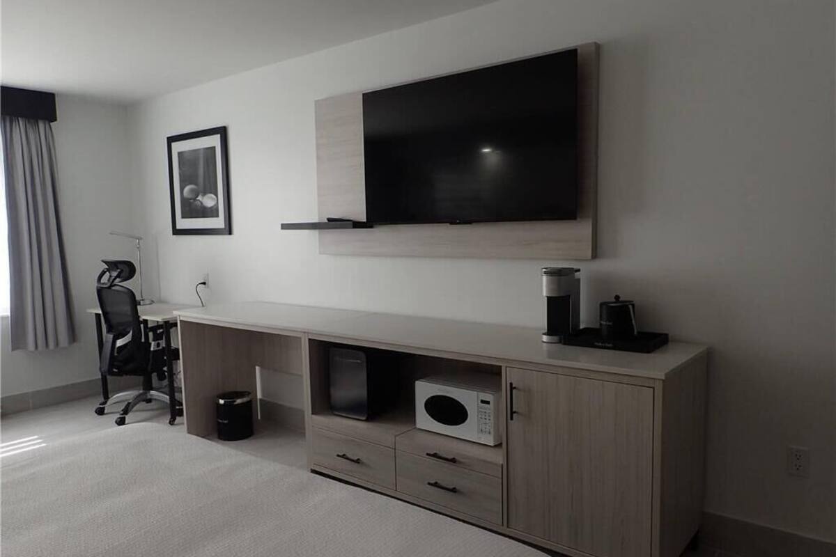 Quarto de hotel moderno com uma grande televisão de tela plana na parede, uma mesa de console de madeira com micro-ondas e uma cadeira de escritório preta.