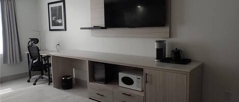 A modern hotel room with a large flat-screen TV mounted on the wall, a wooden console table with a microwave, and a black office chair.