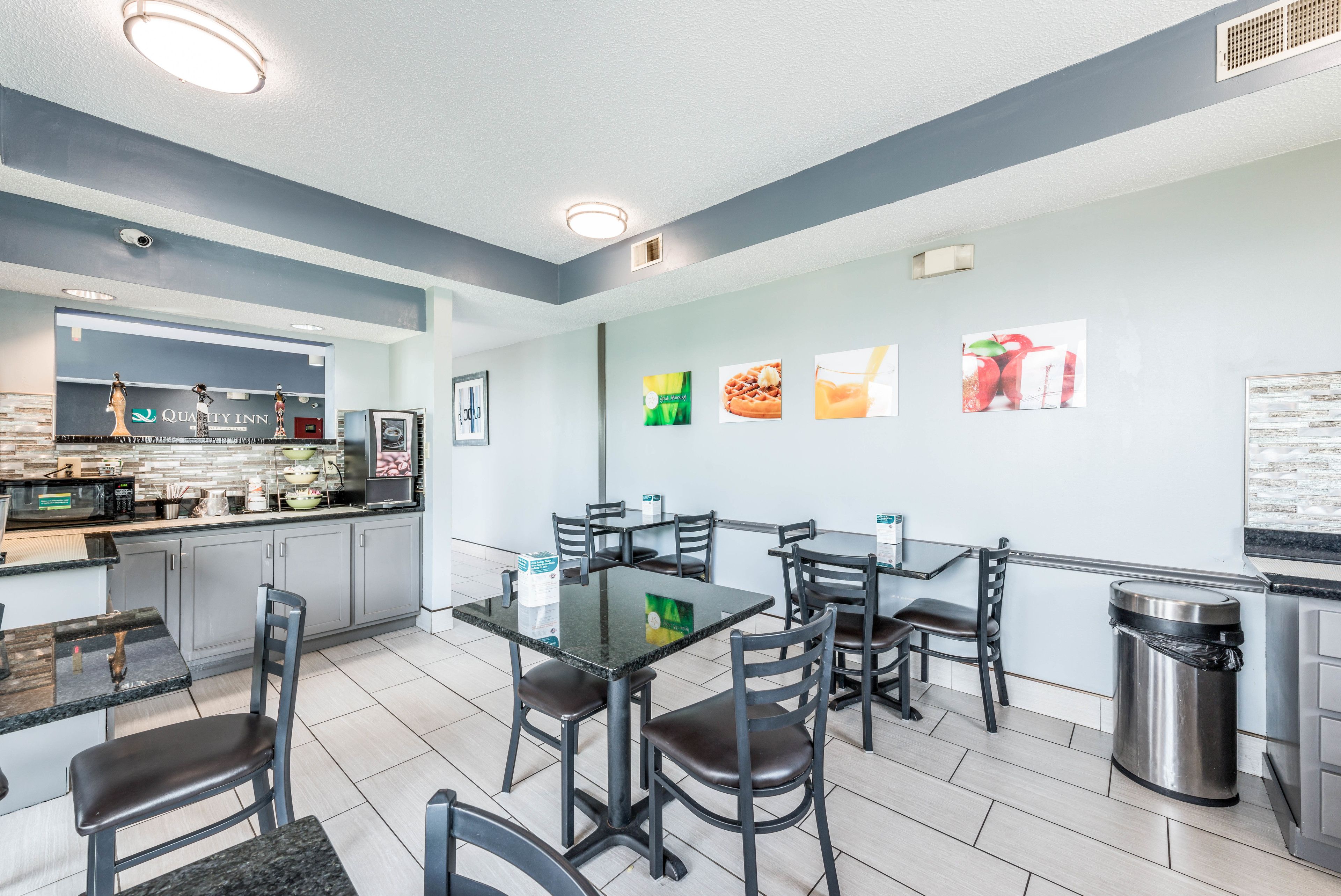 A modern restaurant with a clean, minimalist design, featuring a central dining area with a glass table and black chairs, a kitchen area with a 'Quality Inn' sign, and a wall adorned with food-themed pictures.