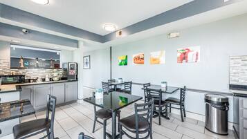 Een modern restaurant met een strak, minimalistisch design, met een centrale eetruimte met een glazen tafel en zwarte stoelen, een keukenruimte met een 'Quality Inn' bord, en een muur versierd met afbeeldingen met daop voedsel thema.
