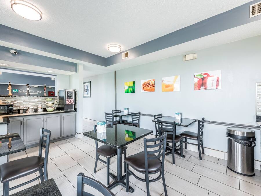 Een modern restaurant met een strak, minimalistisch design, met een centrale eetruimte met een glazen tafel en zwarte stoelen, een keukenruimte met een 'Quality Inn' bord, en een muur versierd met afbeeldingen met daop voedsel thema.