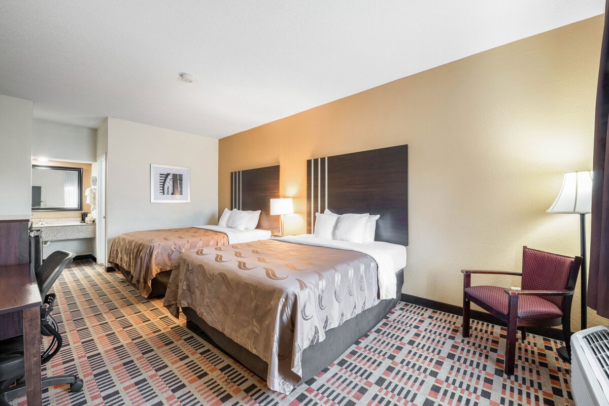 A hotel room with a bed, a desk, a chair, a lamp, and a radiator.